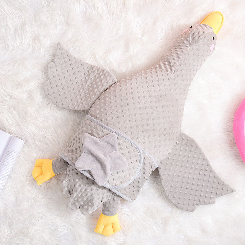 A plush, textured gray goose-shaped blanket with yellow beak and feet is laid on a fluffy white surface. A star-shaped pocket is visible on the body.