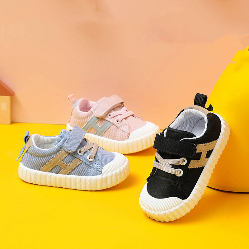 Three colorful toddler sneakers are displayed on a yellow surface. The shoes are in pink, blue, and black, each featuring a strap and a textured white sole.