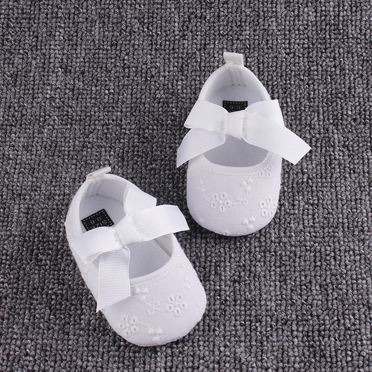 White baby shoes with embroidered floral patterns and large bows on top, set on a textured gray carpet.