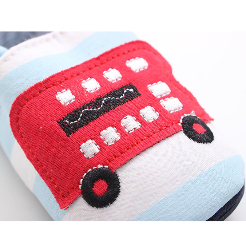 Close-up of a children's fabric shoe with a red double-decker bus applique, featuring white window details and black wheels, set against a light blue and white striped background.