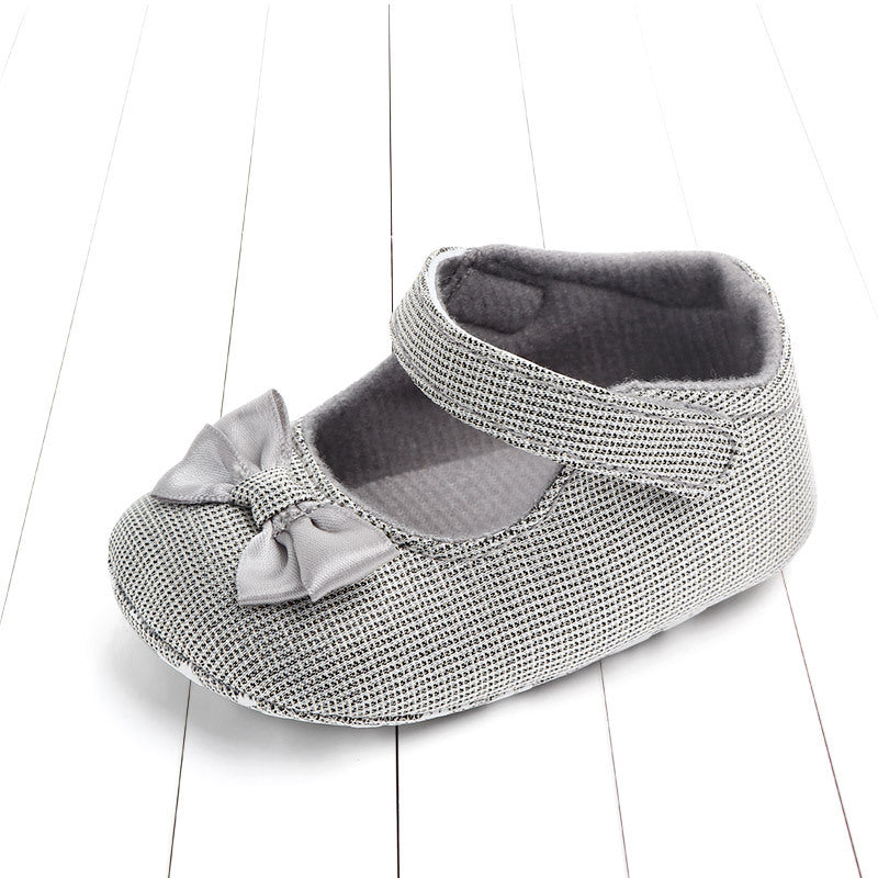 Gray baby shoe with textured fabric, featuring a small bow on the toe and a Velcro strap. It is placed on a white wooden floor.
