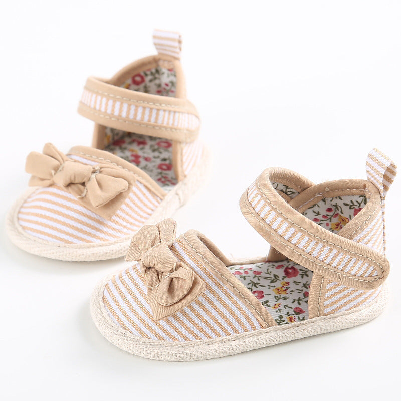 Beige and white striped baby shoes with bows on top, featuring a floral inner lining. They have Velcro straps and soft soles, designed for comfort and style.