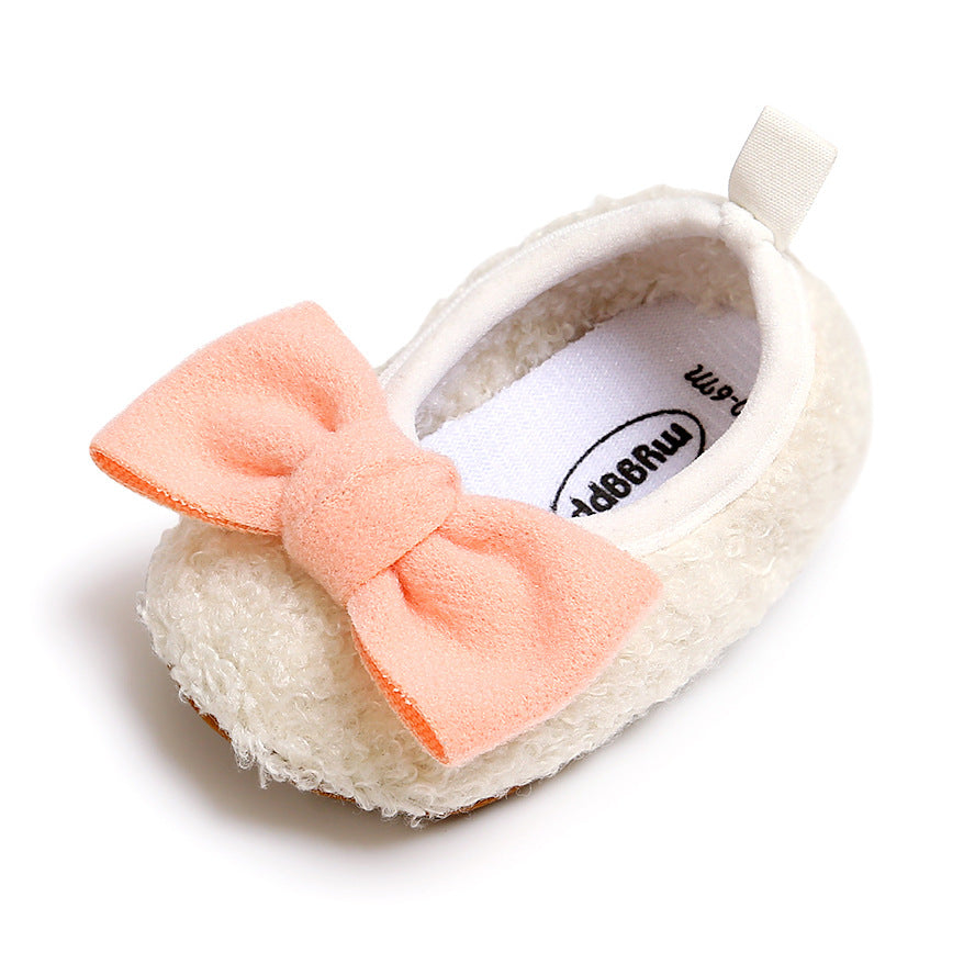 Cream-colored baby shoe with a soft, textured exterior, featuring a large pink bow on top. It has a looped pull tab at the heel and is designed for comfort and style.