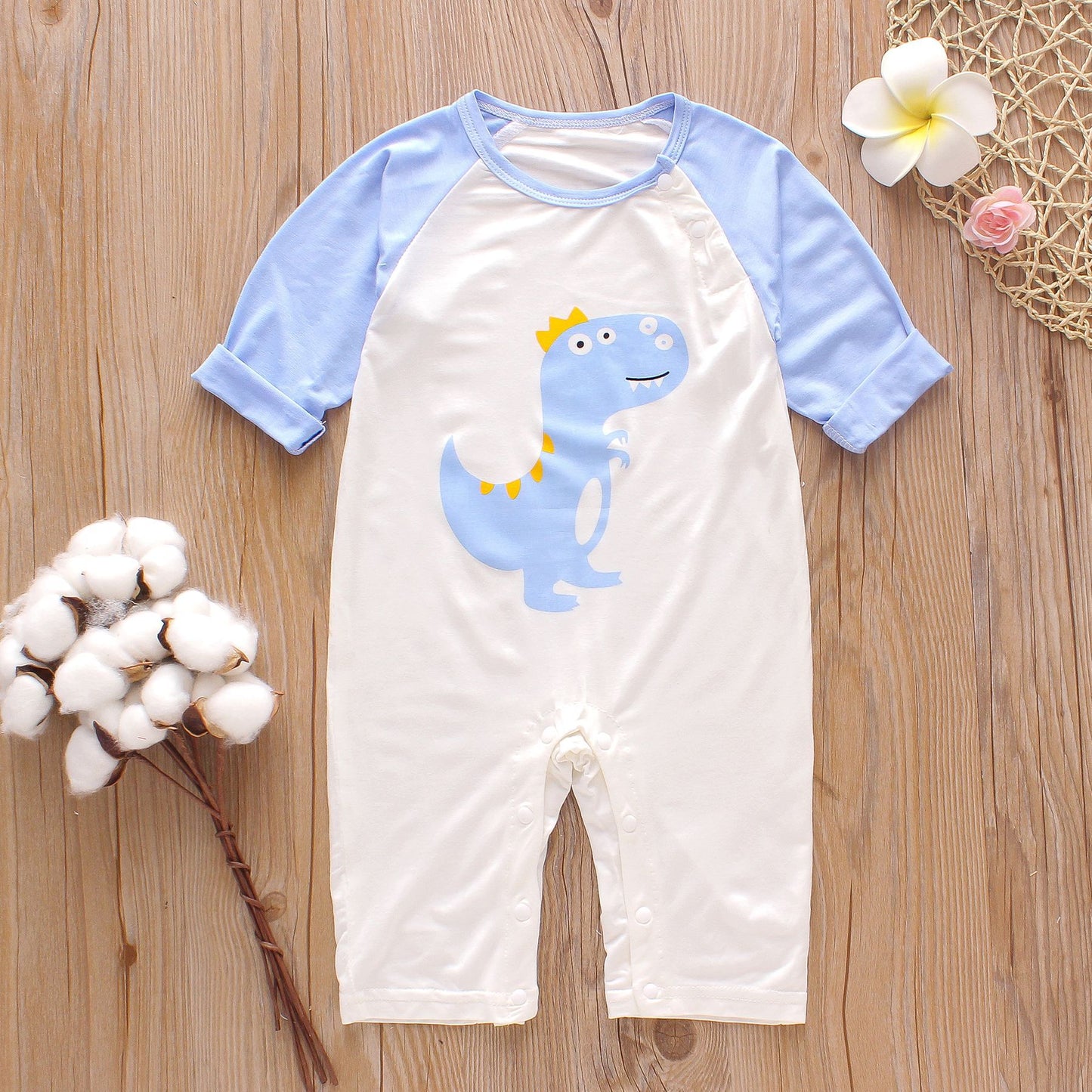 Baby romper with blue sleeves and dinosaur print on a wooden surface. Decorated with cotton branch and flowers.