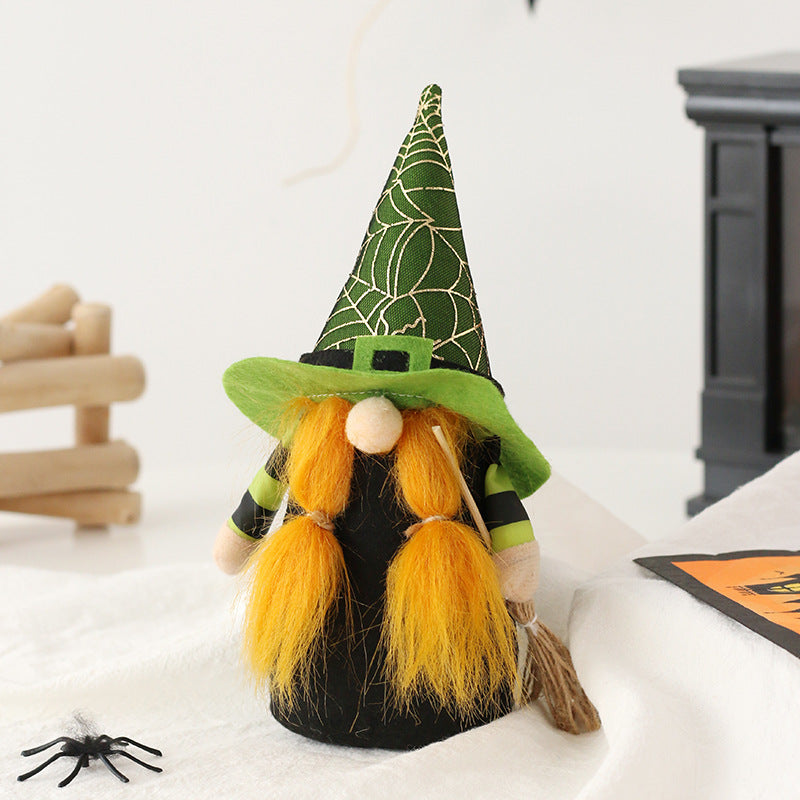 A cute gnome dressed as a witch with a long orange beard and two pigtails, wearing a green hat with a spiderweb pattern. It holds a small broomstick on a white surface. Nearby, a small black spider decoration and wooden logs are visible in the background.