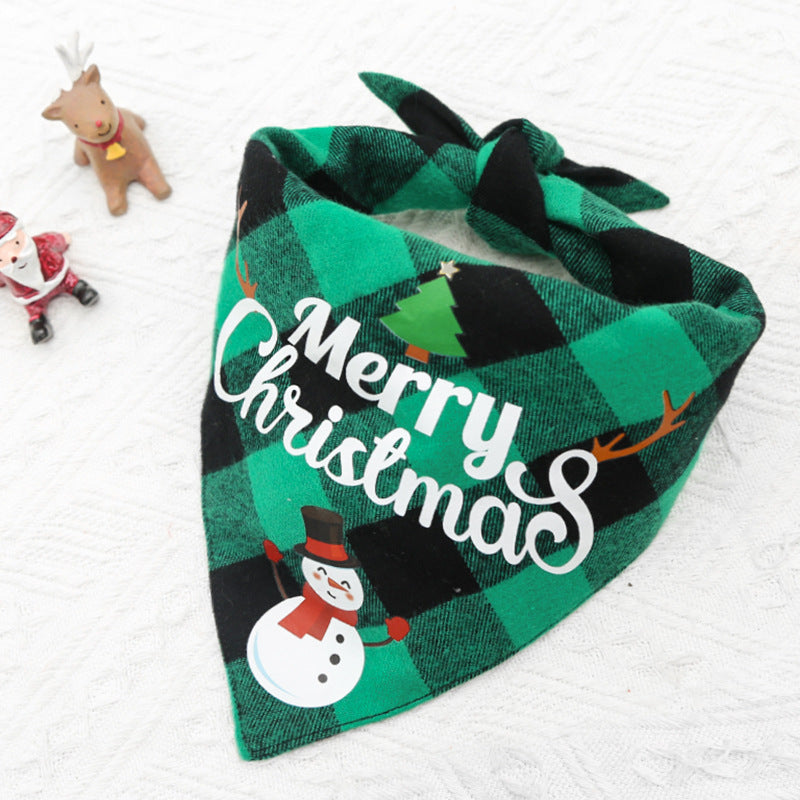 Green and black plaid dog bandana with "Merry Christmas" text, snowman, and tree graphics. In the background, there's a small Santa figurine and a reindeer on a white surface.