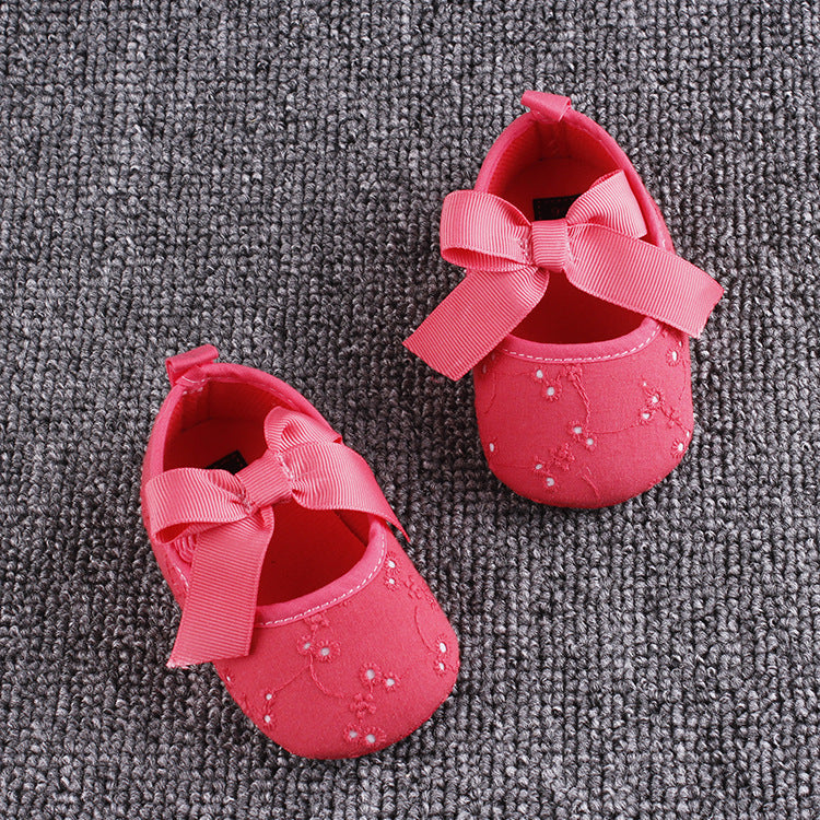 Pink baby shoes with large bows and delicate embroidery, adorned with small beads, placed on a textured gray carpet.