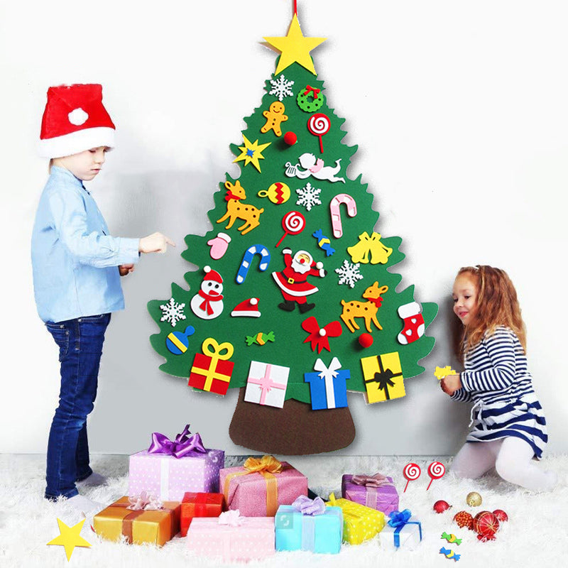 Two children decorate a felt Christmas tree on a wall. The tree has colorful ornaments and is surrounded by wrapped gifts on the floor. One child wears a Santa hat.