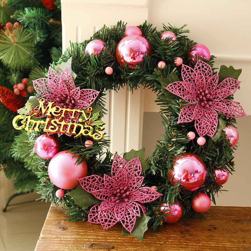 Festive Christmas wreath adorned with pink ornaments, glittery pink poinsettias, and a yellow "Merry Christmas" sign on green foliage, placed on a wooden surface.