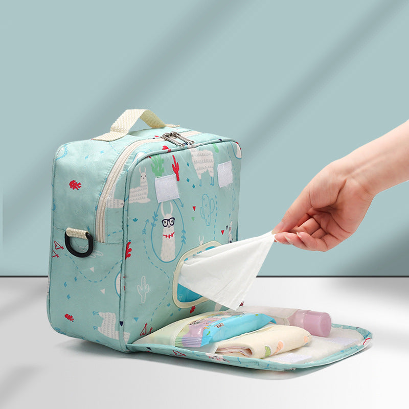 A teal diaper bag with playful illustrations holds baby essentials. A hand pulls a wipe from a built-in dispenser, revealing diapers and a bottle inside an open compartment.