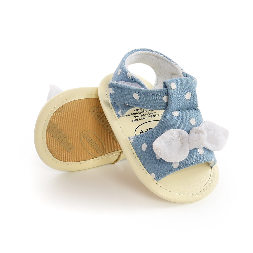 Blue baby sandals with white polka dots, featuring a soft sole and a decorative white bow on the front. One sandal is upright while the other is tilted, showing the sole.