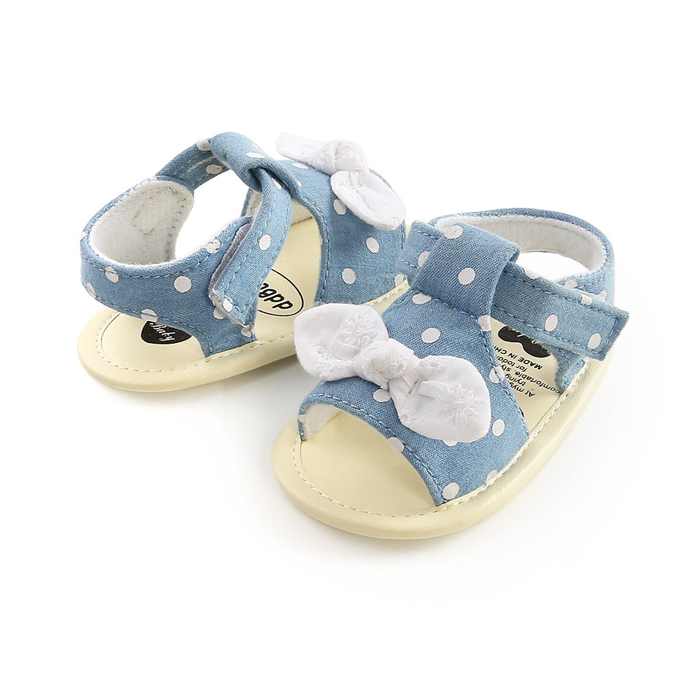 Blue polka dot baby sandals with white bows, featuring Velcro straps and soft sole, angled view.