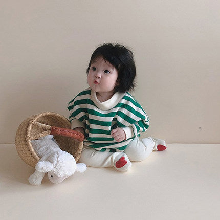 A baby sits on the floor wearing a green and white striped sweater, with white pants and red socks. The baby looks curious, next to a tipped basket holding a plush lamb.