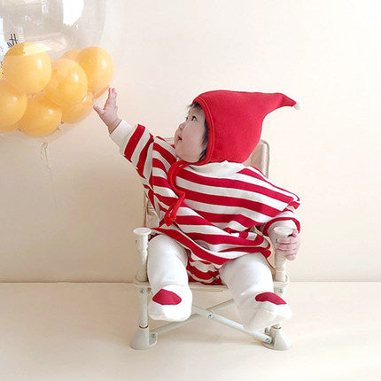 A baby in a red and white striped outfit with a red hat sits on a small chair, reaching towards a cluster of yellow balloons floating nearby against a plain background.