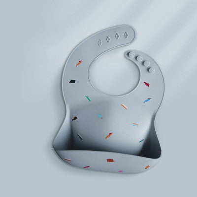 A gray silicone bib with a deep pocket and colorful geometric shapes. Fastens with buttons on the side, designed to catch food spills, shown against a light background.