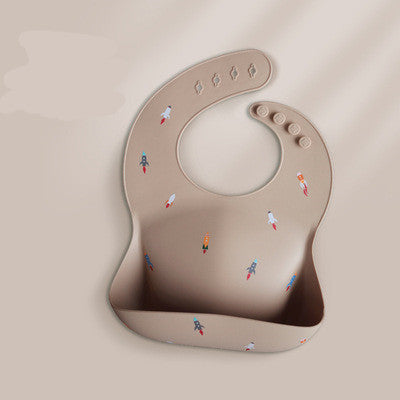 A beige silicone baby bib with colorful rocket patterns, featuring adjustable button closures and a wide pocket for catching food, laid flat on a light background.