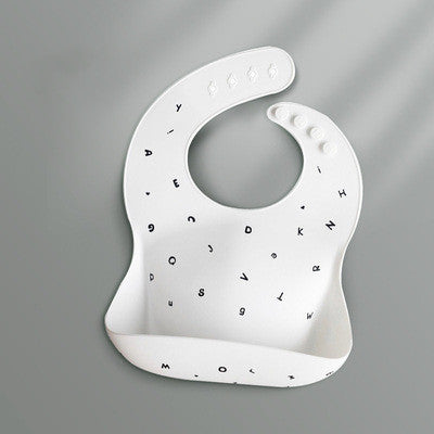 A white silicone baby bib with scattered black letters, featuring a pouch at the bottom for food-catching. It has an adjustable neck clasp with three buttons, set against a gray background.