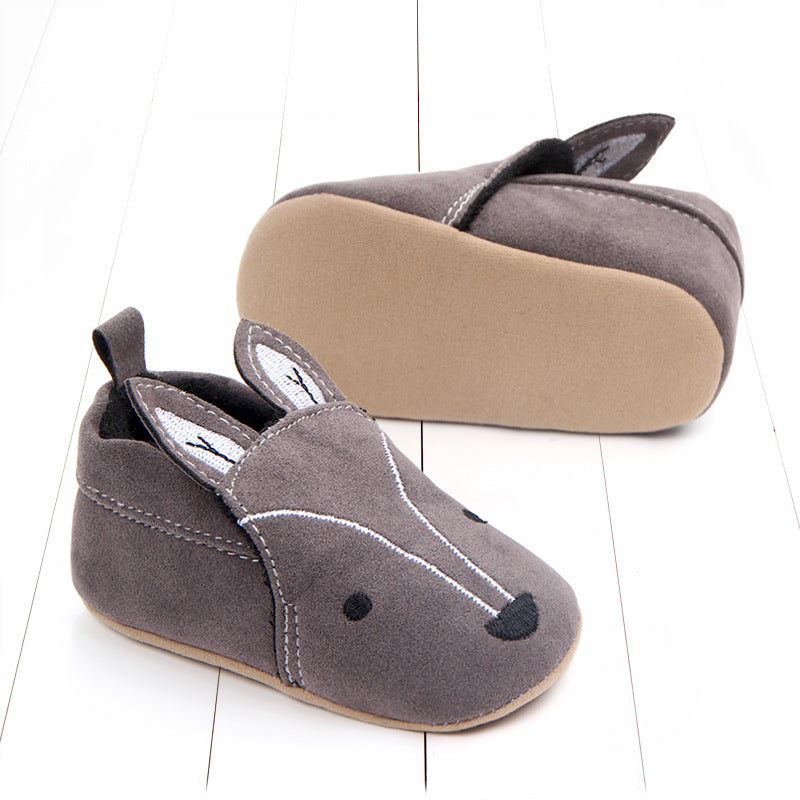 Gray baby shoes with bunny ear design, featuring stitched eyes and nose. One shoe is upright, while the other shows the sole. Set on a white wooden background.