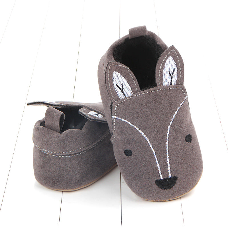 Gray baby moccasins with fox face design and cute ear details on white wooden floor. Front shoe stands upright while rear shoe lies on its side.