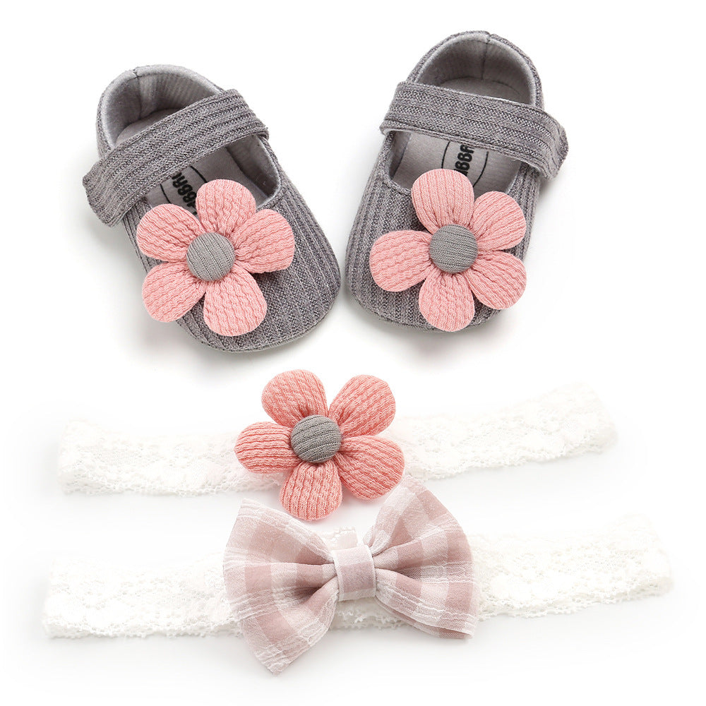 Gray baby shoes with pink flower decorations, alongside two lace headbands, one with a pink flower and the other with a checkered bow.