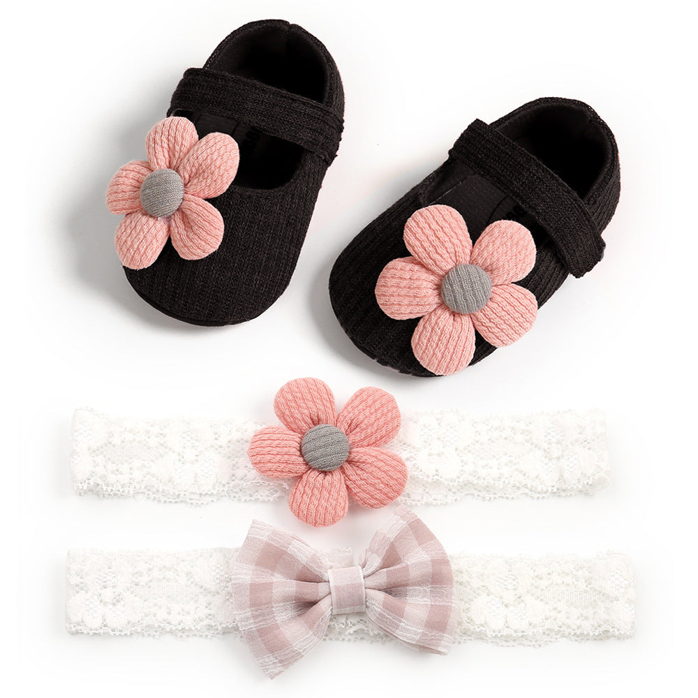 Black baby shoes with pink flower embellishments, paired with two white lace headbands—one with a pink flower and the other with a checkered bow.