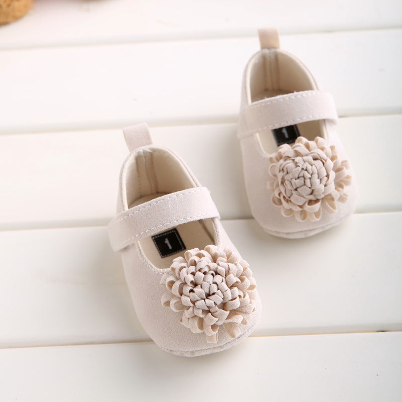 Cream-colored baby shoes with a floral embellishment on the toes, featuring a strap for closure, displayed on a white wood surface.
