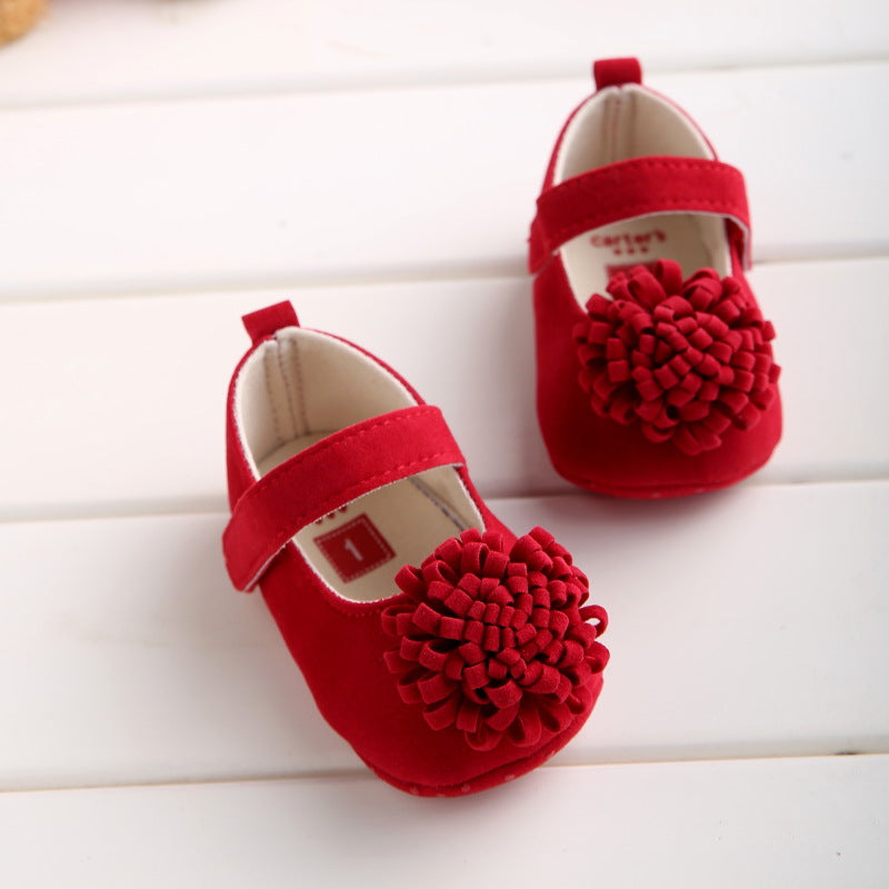 Red baby shoes with a front flower embellishment, velcro strap, and soft interior. Placed on a white wooden surface, they're ideal for special occasions or everyday wear.