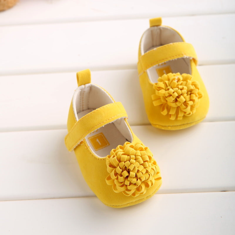 Yellow baby shoes with Velcro straps and pom-pom decorations on the toes, placed on a white wooden surface.