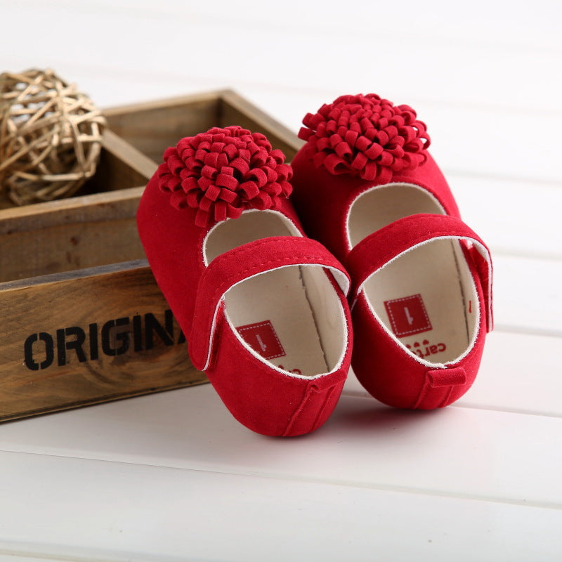 Red baby shoes with textured flower decorations on the toes, set on a white surface. Background includes a wooden box and decorative rattan ball.
