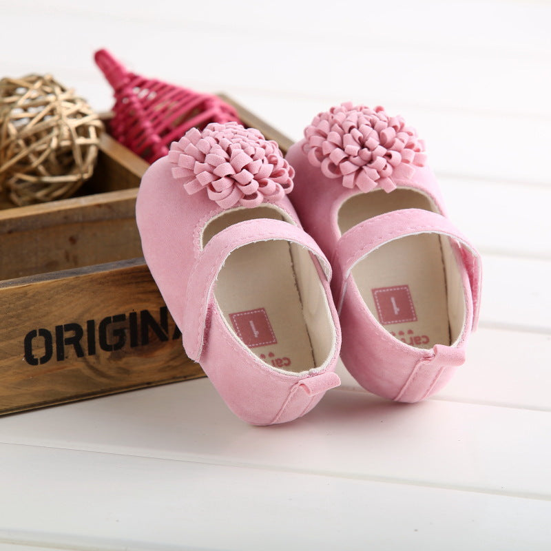 Pink baby shoes with large flower appliques on top, placed on a wooden crate labeled "ORIGINAL," with decorative wicker and red items in the background on a white surface.