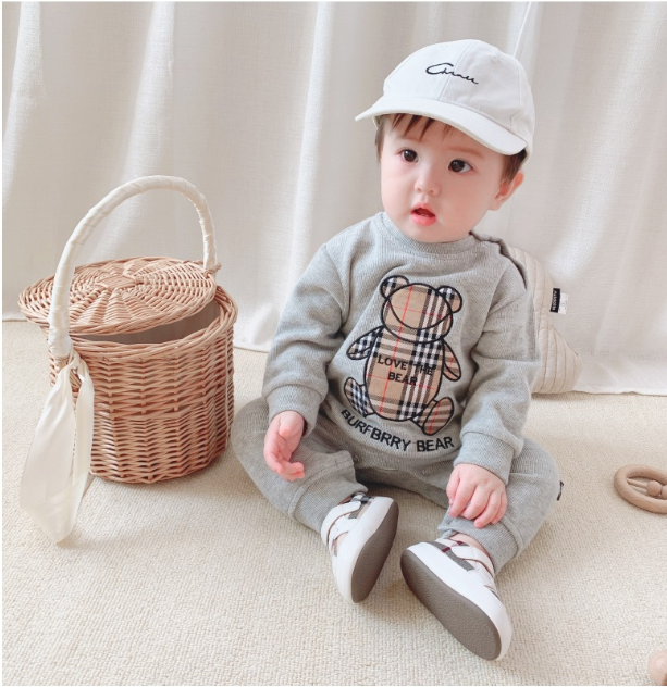 A baby in a gray outfit with a teddy bear design sits on the floor. The baby wears a white cap and sneakers. A wicker basket with a cloth rests beside them.