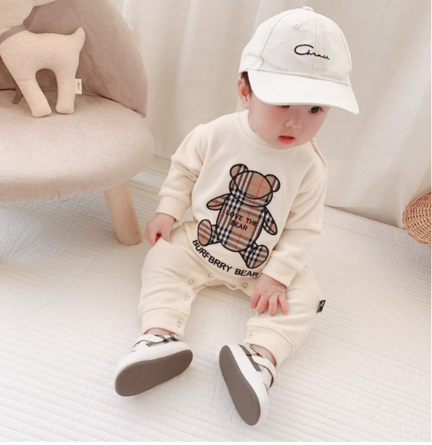 A toddler wearing a matching beige outfit with a plaid bear design, white sneakers, and a baseball cap sits on a white surface. A plush deer toy is in the background.