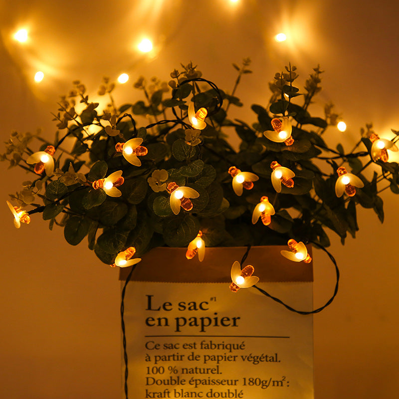 A paper bag labeled "Le sac en papier" holds a plant adorned with glowing firefly fairy lights, set against a blurred backdrop of warm, ambient lights.