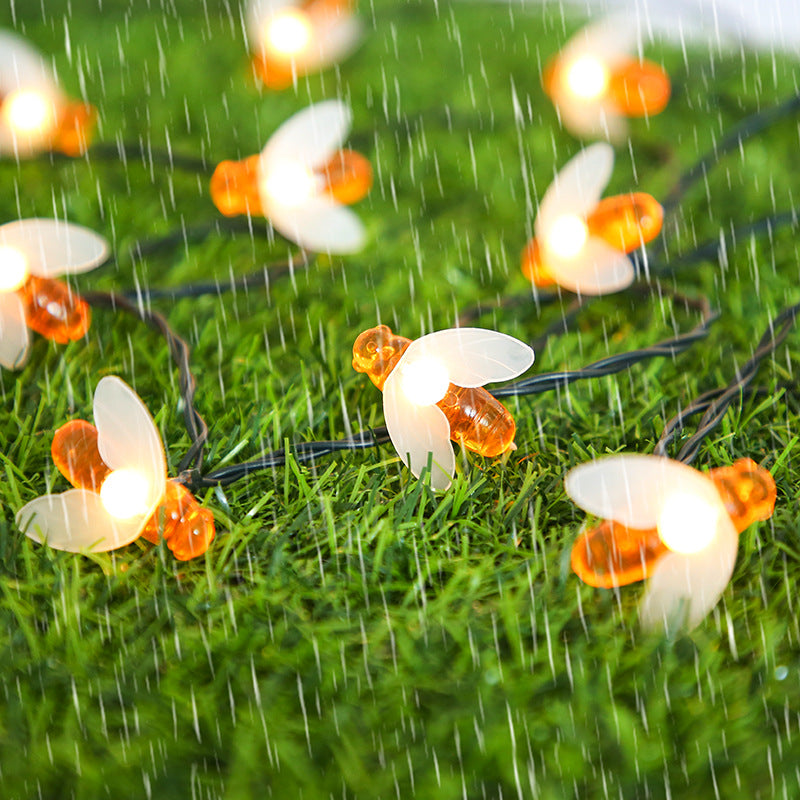 Glowing bee-shaped string lights on wet grass, illuminated against a backdrop of light rain creating a cozy outdoor atmosphere.