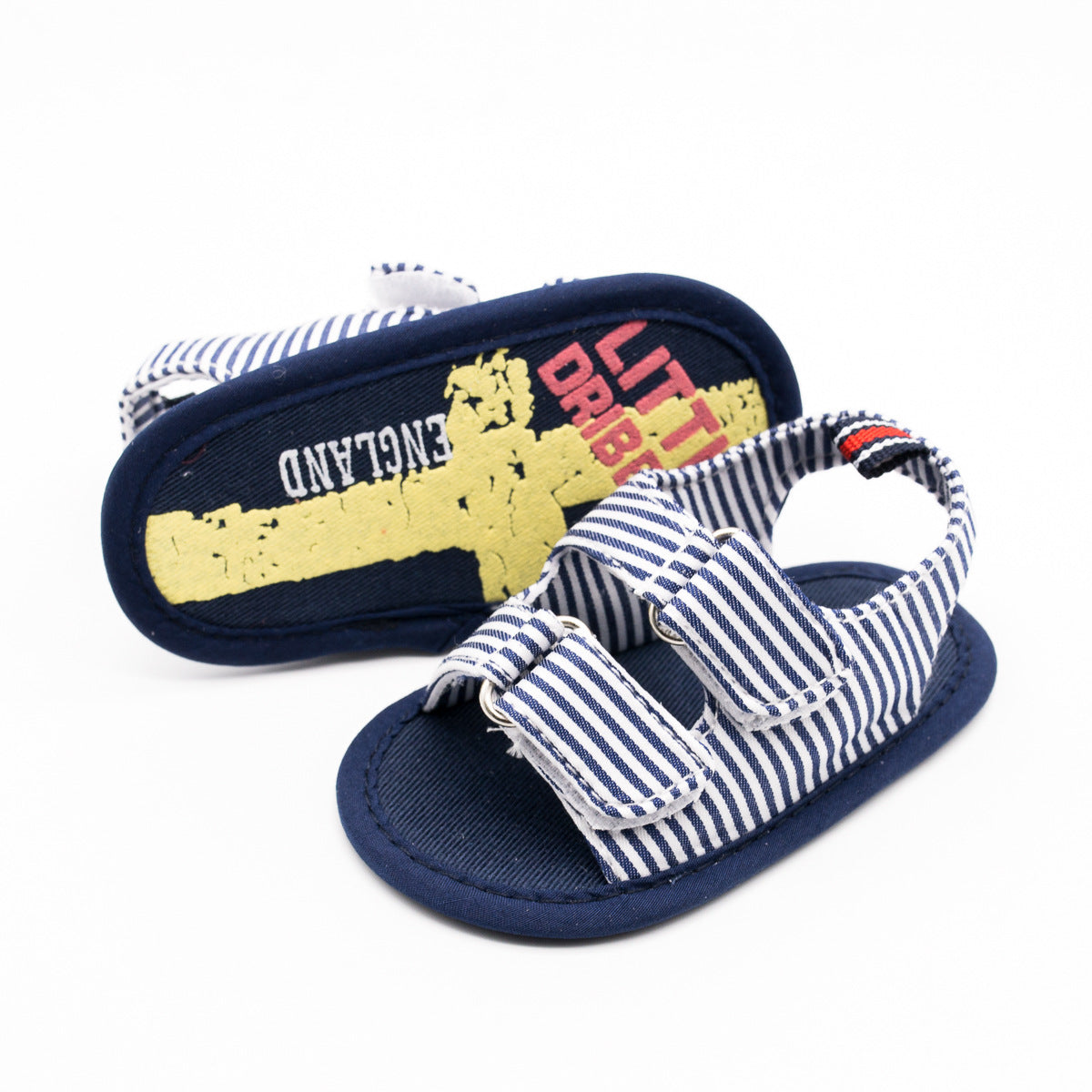 Striped baby sandals with blue and white straps. One shoe shows the sole with "LITTLE DRIBBLER ENGLAND" text in red and white over a yellow graphic.