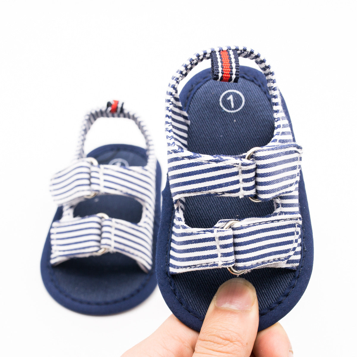 Close-up of blue and white baby sandals with striped straps. One sandal is held in a hand, displaying its soft sole and adjustable buckle design.