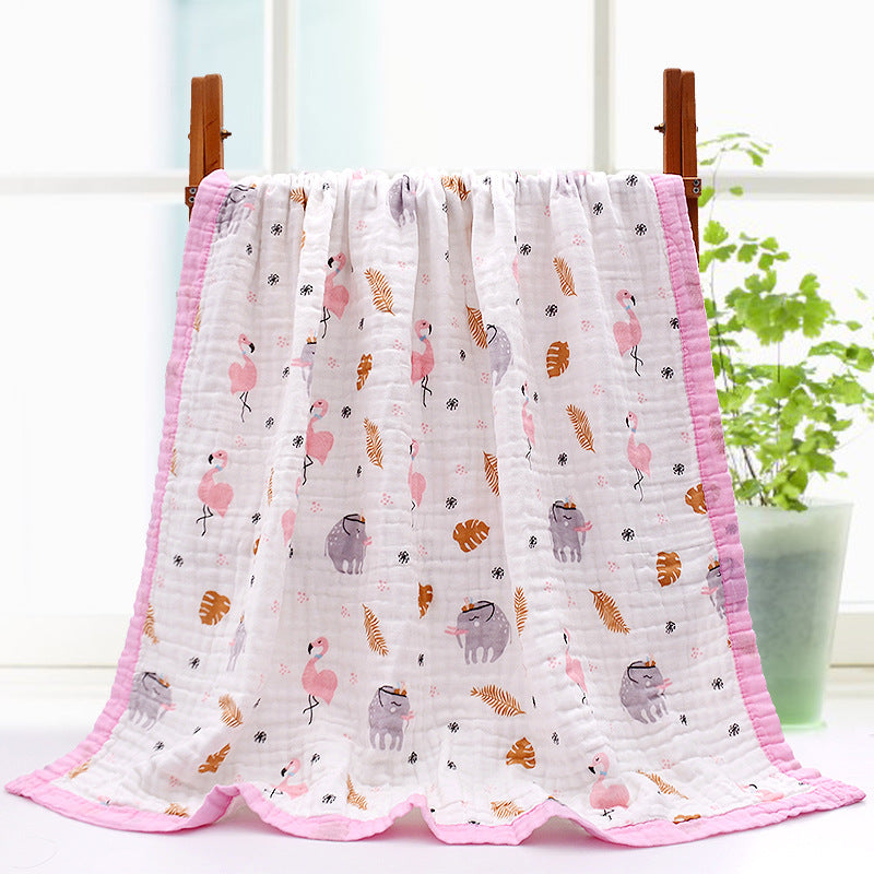 A soft baby blanket draped over a stand features a pink border and playful prints of flamingos, sloths, leaves, and flowers in pastel colors, set in a bright room with a potted plant.