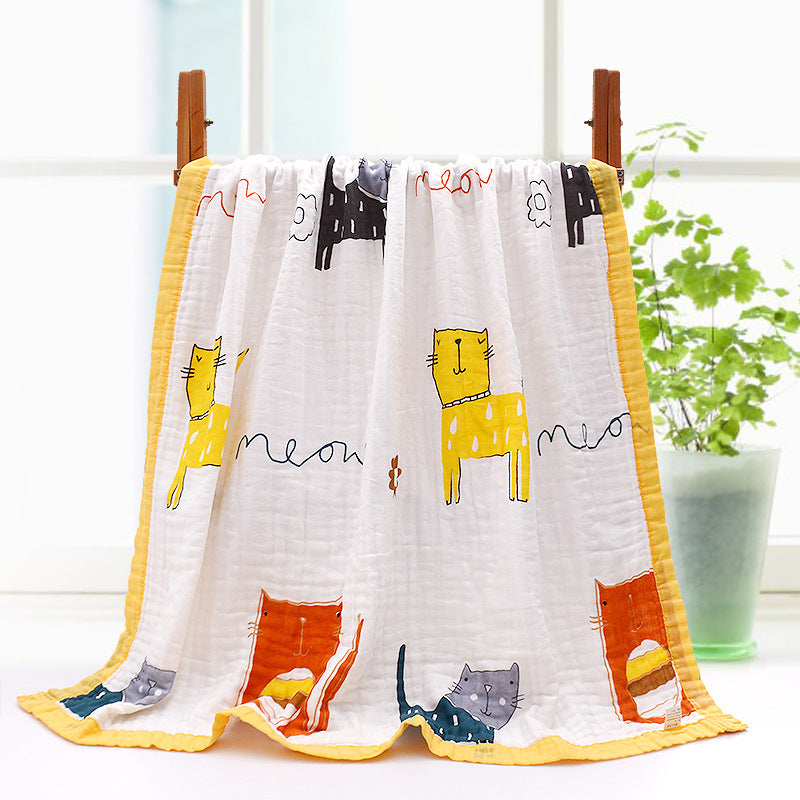 A colorful baby blanket with playful cat illustrations and "meow" text in various styles, hung on a wooden rack, bright yellow edges, with a potted plant in the background.