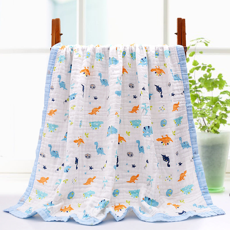 A soft, white baby blanket with colorful dinosaur prints in blue, orange, and green. It is draped on a wooden rack by a window, with a light blue border. A plant is in the background.