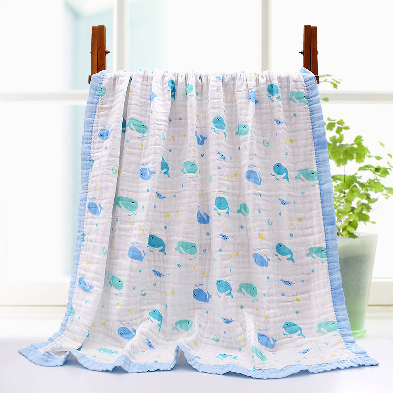 A soft, white quilt with blue trim is draped over a wooden hanger, featuring cheerful blue and green whale patterns. A potted plant sits nearby, with sunlight filtering in.