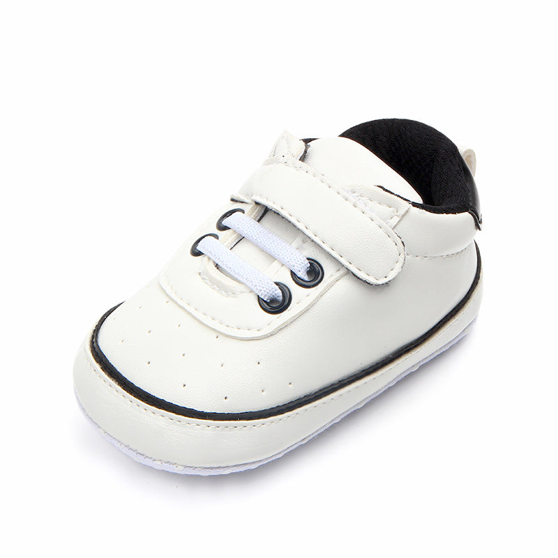 White baby shoe with a black sole and details. Features white laces and a Velcro strap for easy fastening. The shoe has small perforations for breathability.