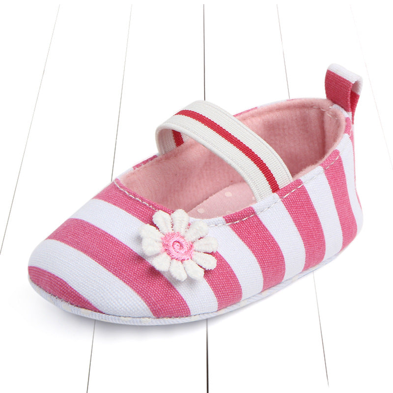 Pink and white striped baby shoe with a white flower and pink center. It has a matching striped elastic strap and a soft, pink interior, positioned on a white wooden surface.