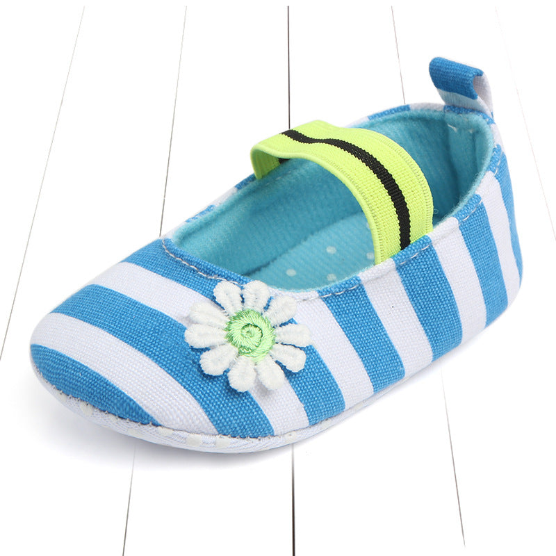 Blue and white striped baby shoe with a decorative white flower on the side and a green elastic strap, placed on a white background with light slats.