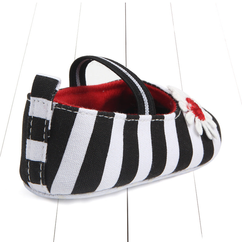 Black and white striped baby shoe with red interior, a white flower with a red center on top, and a thin black strap over the foot, placed on a white wooden floor.