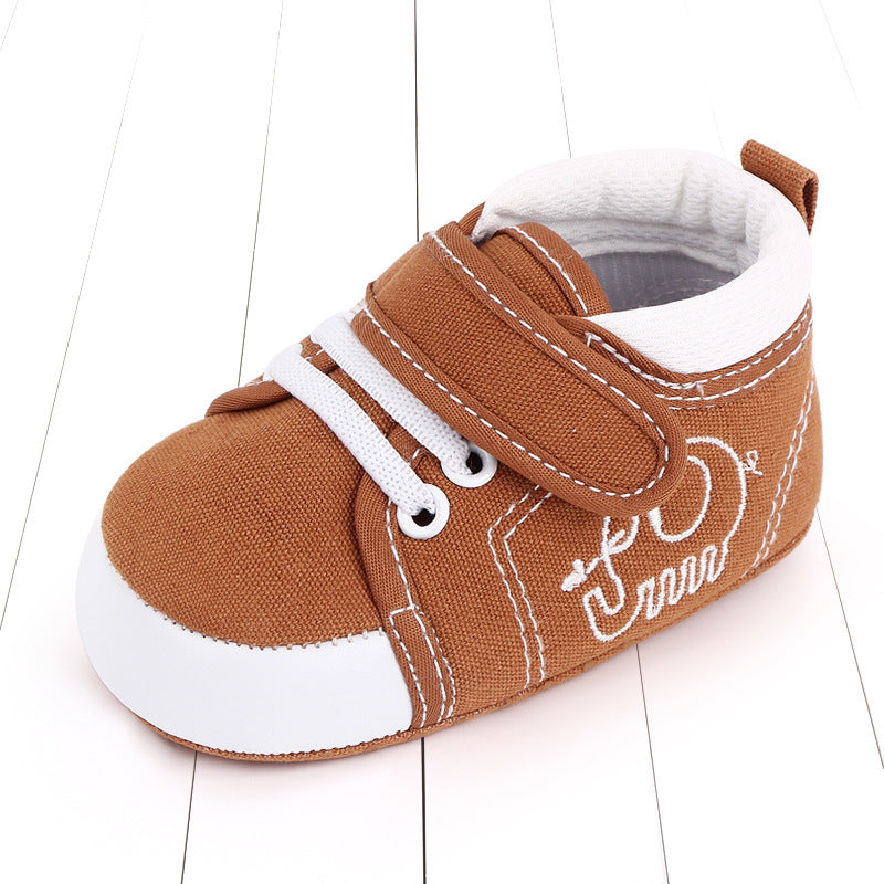 Brown baby shoe with white accents on a wooden floor. Features a Velcro strap, white laces, and an embroidered design on the side.