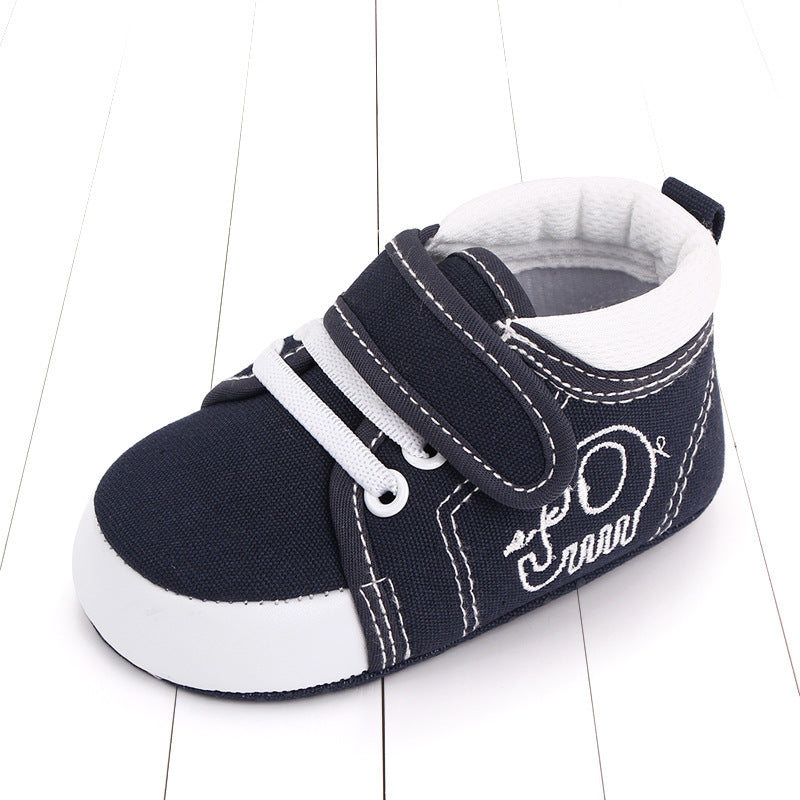 A navy blue baby shoe with white accents and laces, featuring a Velcro strap and embroidered design, placed on a white wood surface.