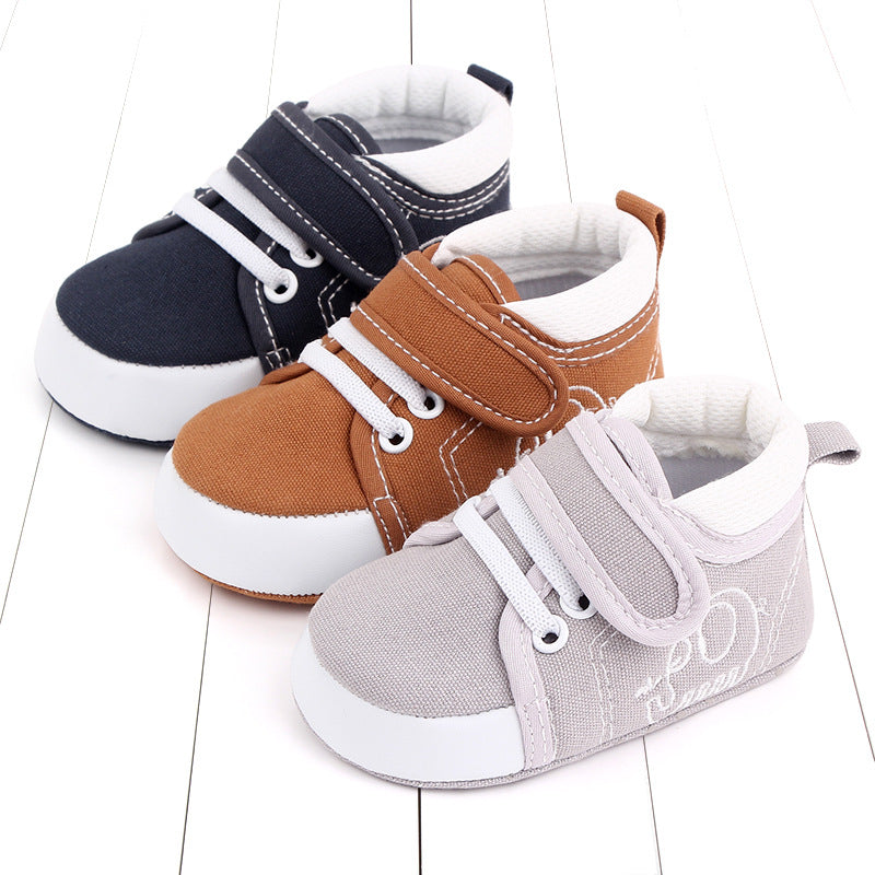 Three pairs of baby shoes on a white wooden floor. They are dark blue, brown, and light gray with white laces and velcro straps, featuring a casual sneaker design.