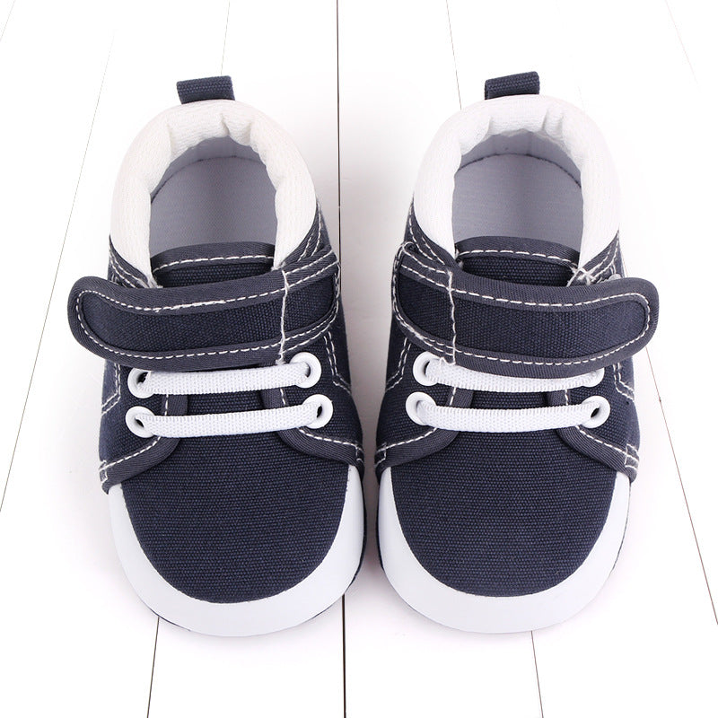 Navy blue infant shoes with white soles and laces. They feature velcro straps and contrasting stitching, positioned side by side on a white wooden surface.