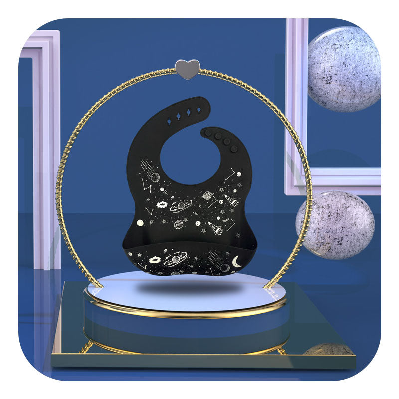 A black bib with space-themed illustrations is suspended within a gold circular stand. The background is dark blue with decorative frames and spherical elements.