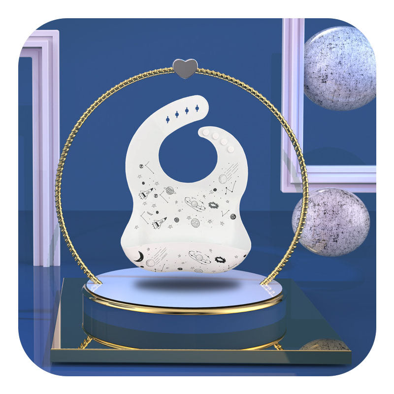 A white baby bib adorned with space-themed illustrations is displayed on a rotating platform, framed by a gold circular stand with a heart accent, set against a blue background.